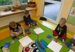 Troje dzieci siedzi na dywanie przed płytką i z ułożonymi klockami w szeregu według wzrastającej wartości. Dzieci przeliczają oczka na poszczególnych klockach.